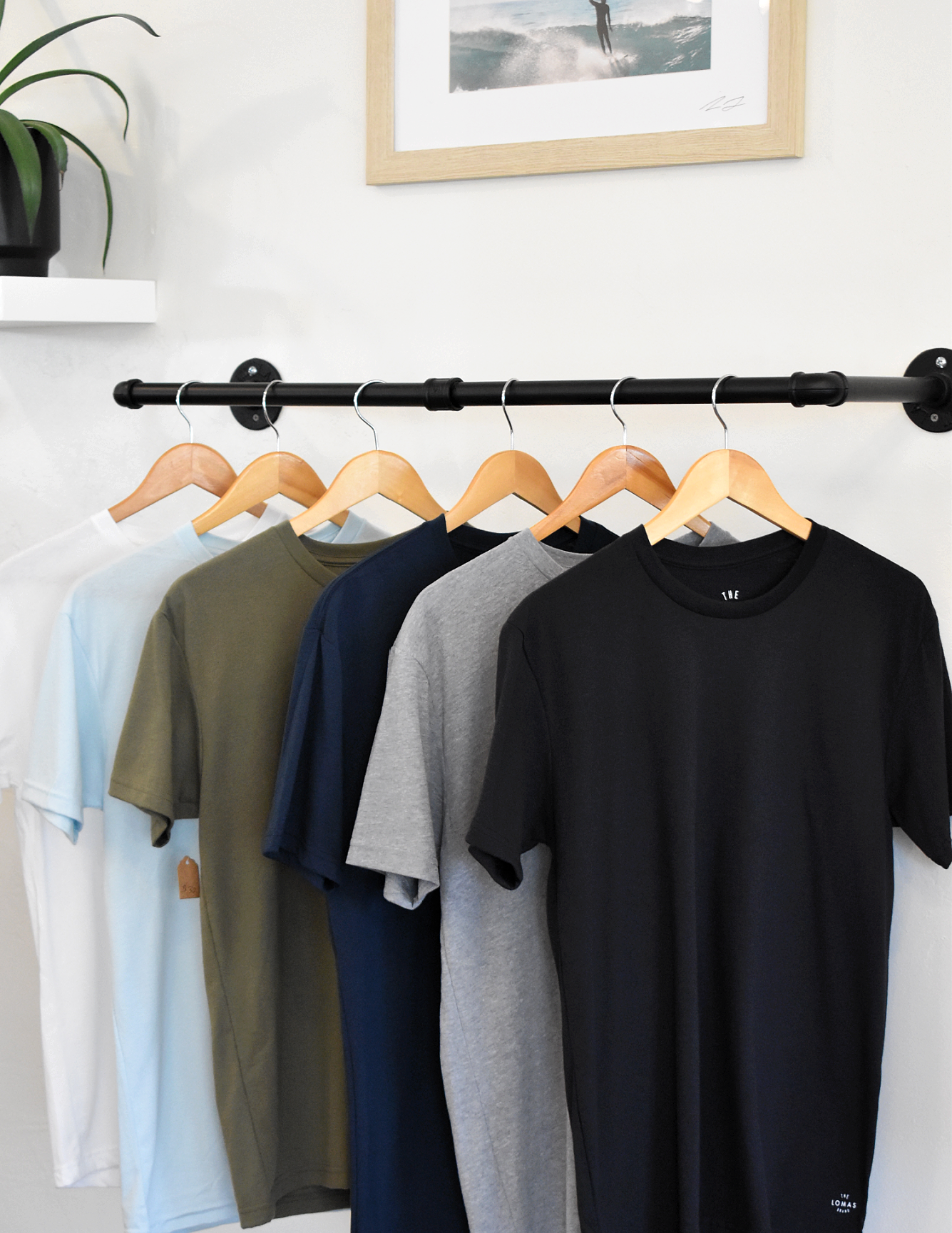 Assorted colored shirts (white, blue, army green, navy, grey, and black) hanging on wooden hangers. A framed photo and a plant are in the background.
