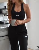 Mid body photo of a woman wearing black joggers and a black sports bra. Both pieces feature the same white logo with a cactus, a bird, and a rising sun. She is holding a coffee mug.