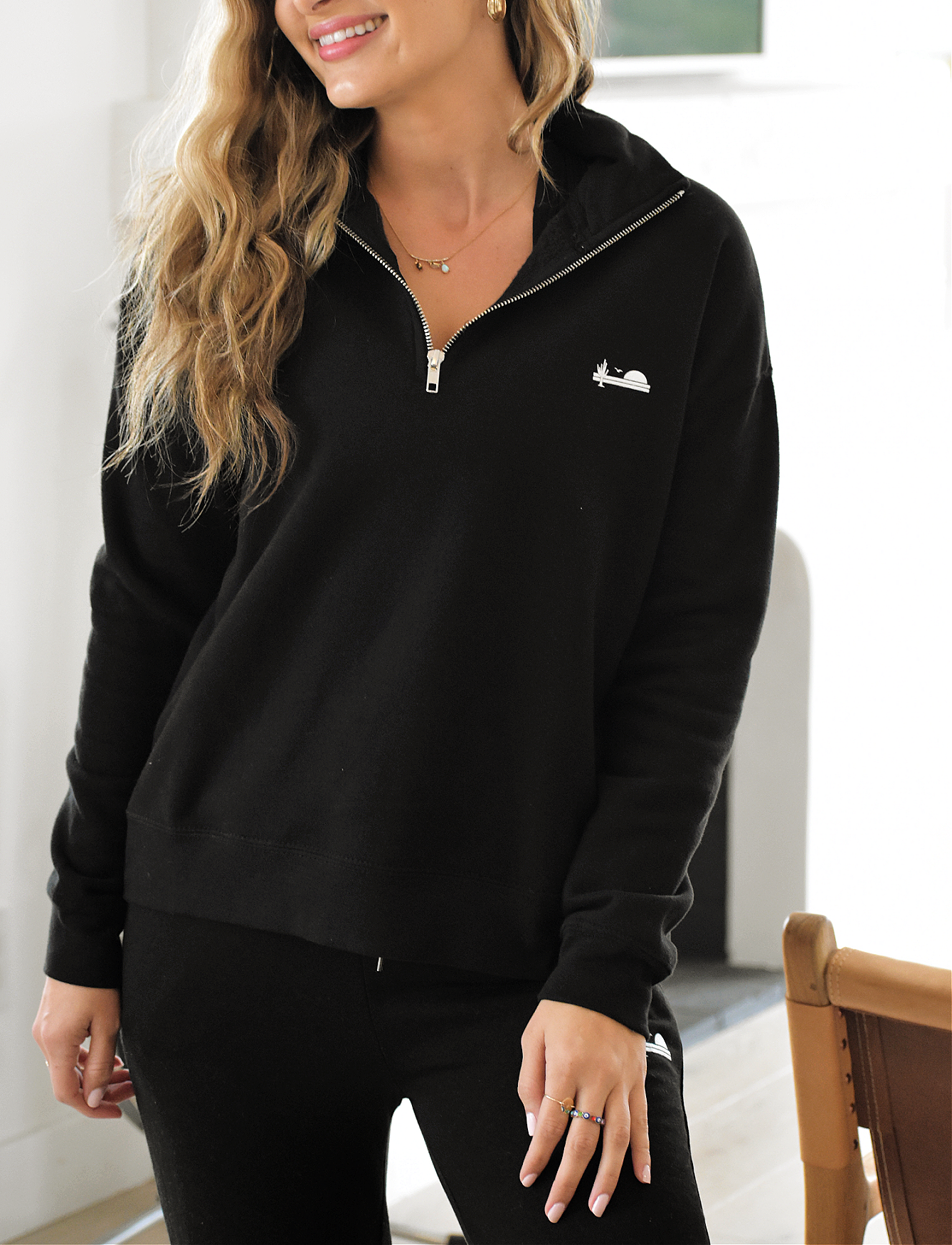 A woman wearing a black quarter-zip sweatshirt with a small white logo on the chest. The sweatshirt is partially unzipped, revealing a necklace. She has long, wavy blonde hair.
