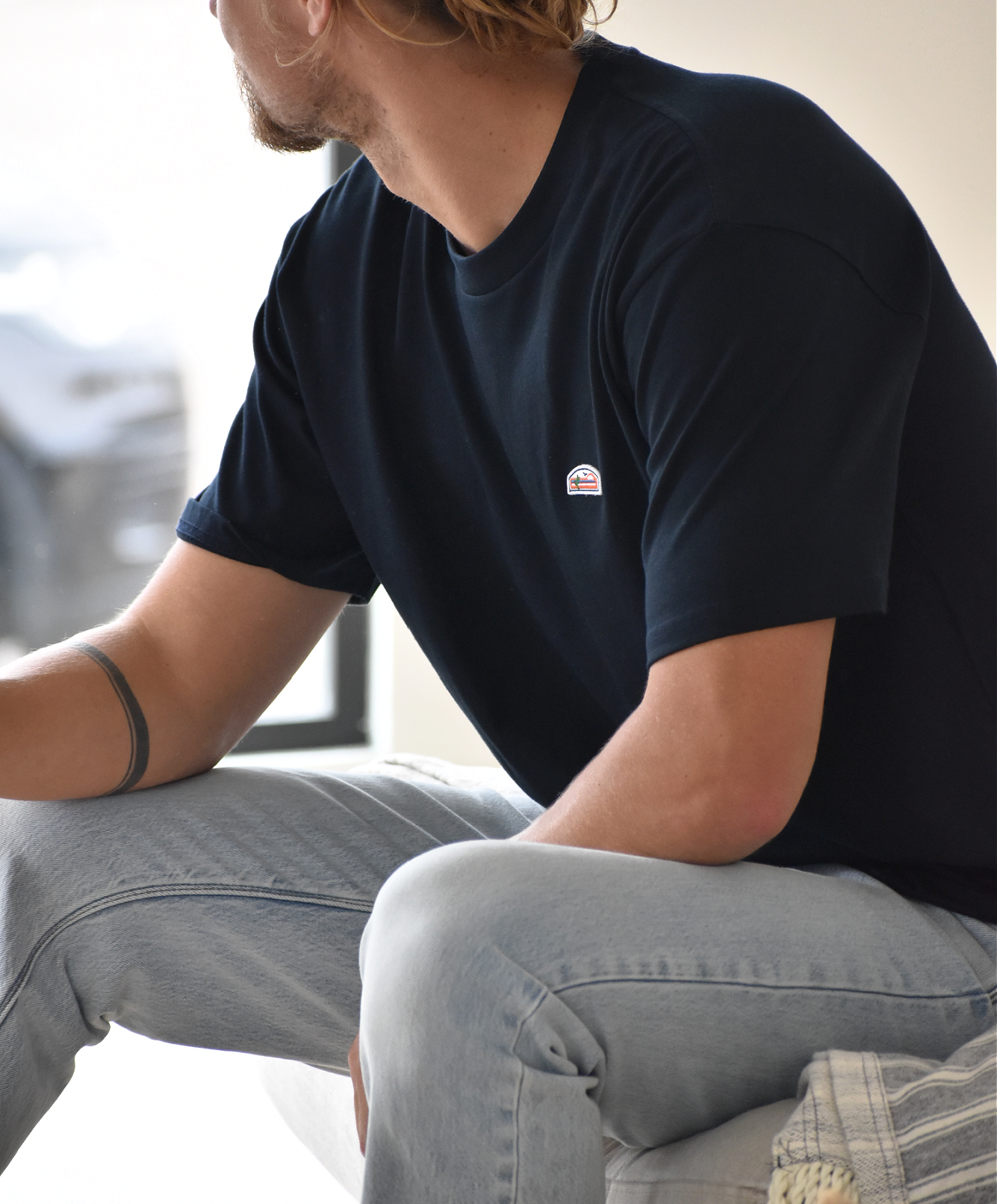 A close-up of a man's head and upper back, showing his hair tied in a bun and the back of his navy t-shirt with a small green, orange, and white logo patch visible on the upper right chest.