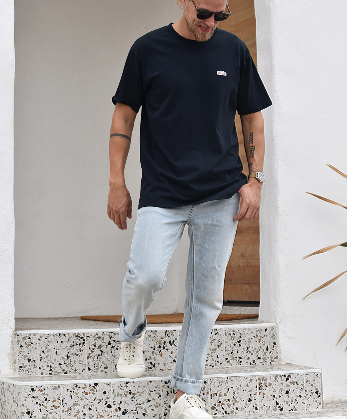 A man standing on what appears to be terrazzo steps, wearing a navy t-shirt and light blue jeans. The shirt has a small green, orange, and white logo patch visible on the upper right chest. He's looking down and smiling.