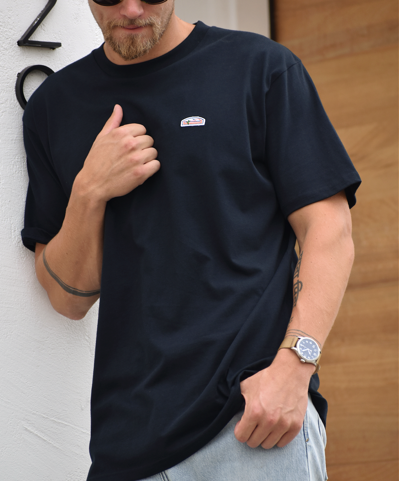 A close view of a man's upper body, showing his beard, sunglasses, and a navy t-shirt. The shirt has a small green, orange, and white logo patch visible on the upper right chest. The man is making a gesture with his hand near his chest.