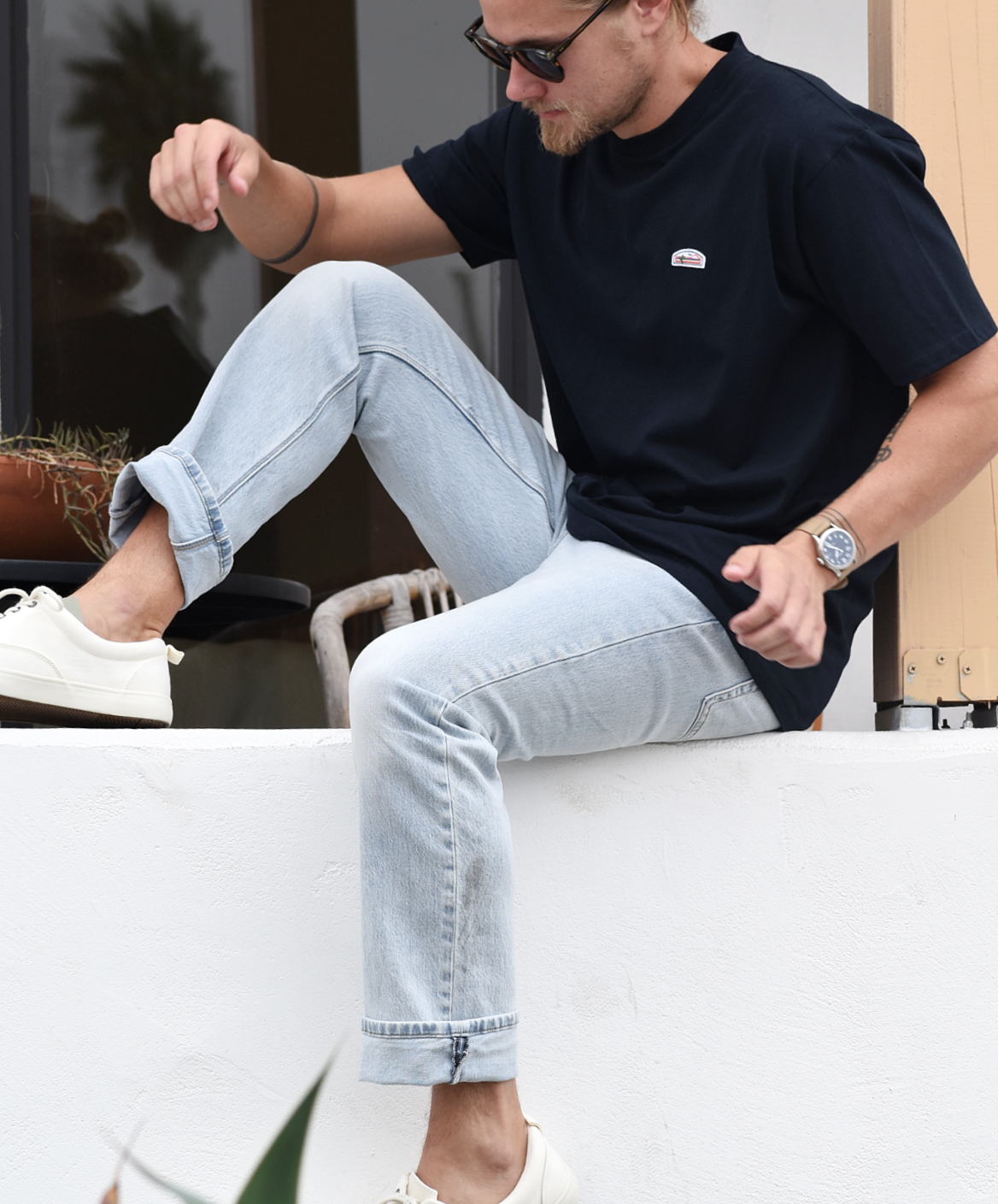 A man sitting on a ledge or bench, wearing a navy blue t-shirt with a small orange, green, and white logo patch on the chest, light blue jeans, and white sneakers. He has long blonde hair tied back and is wearing sunglasses.