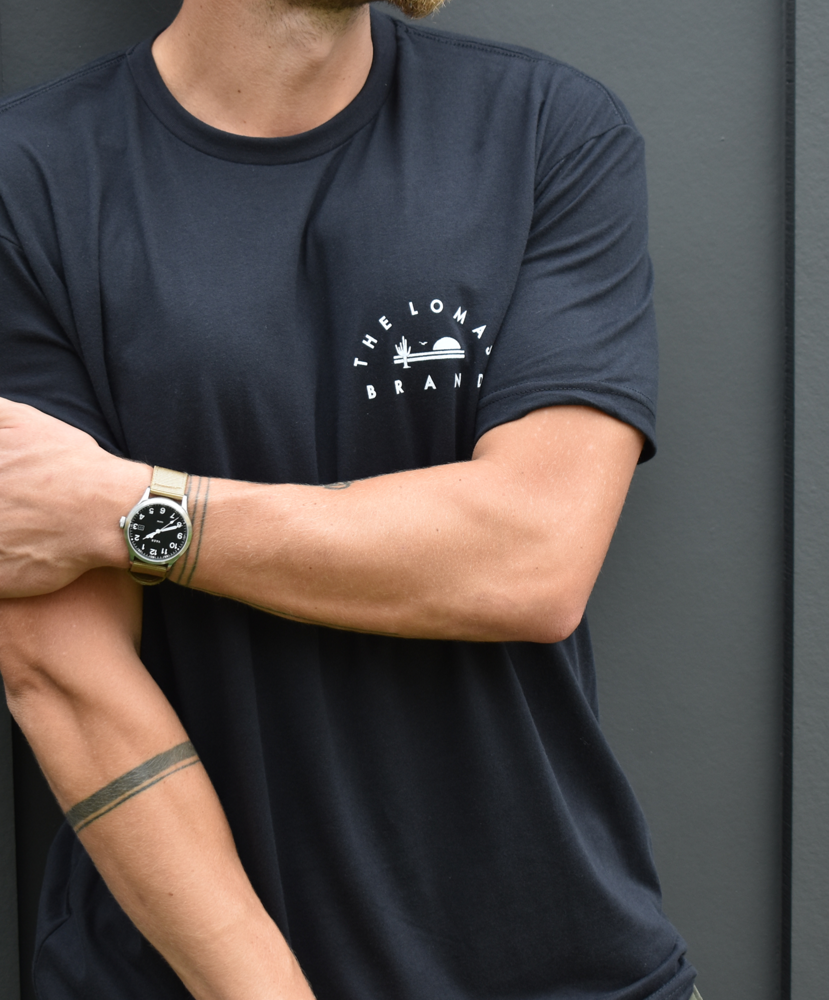 Man wearing a black t-shirt with "THE LOMAS BRAND" logo on the chest. The logo features a sun setting behind desert mountains and a cactus. The person's face is not visible, only their torso and crossed arms.