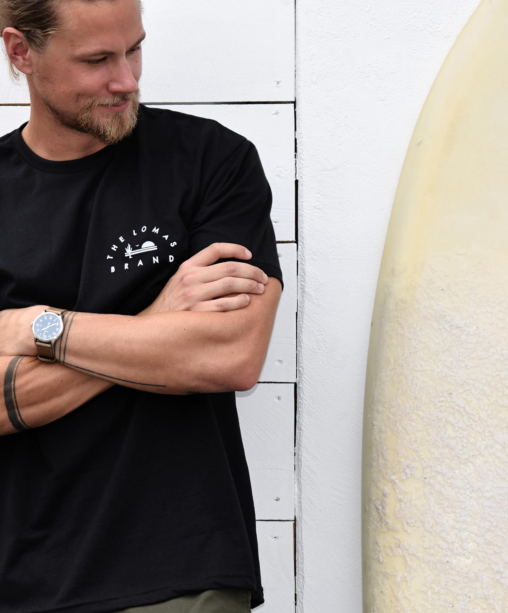 Man wearing a black t-shirt with a small white logo on the chest. The logo says "THE LOMAS BRAND" in a circular font around a saguaro cactus, a bird, and a rising sun. He is standing next to a surfboard, has blonde hair and a beard, and is smiling slightly while looking to the side.