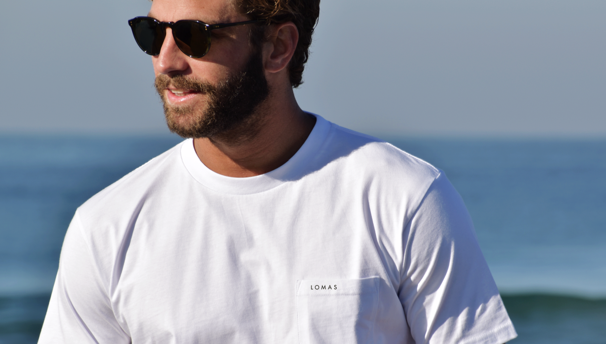 Man with sunglasses staring off camera wearing a crisp yet soft white pocket t-shirt that says 'Lomas' in all caps on the seam. The background is the ocean. 