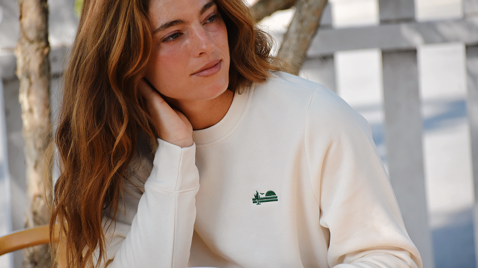 Woman with short dark hair wearing a grey crewneck sweatshirt with the sleeves pulled up. The sweatshirt features a small patch with The Lomas Brand logo on the upper left chest. The woman looks to the side and has her hands on her hips. 