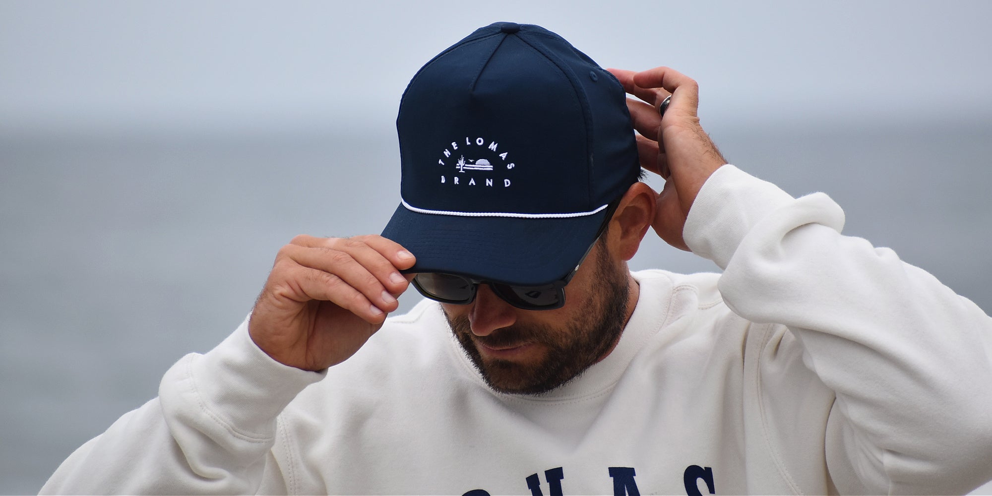 High contrast photo of a man fixing the brim of his navy blue hat with The Lomas Brand's colorful patch on the front. The man is wearing a white shirt and carrying a black jacket. The backdrop is the blue sky. 