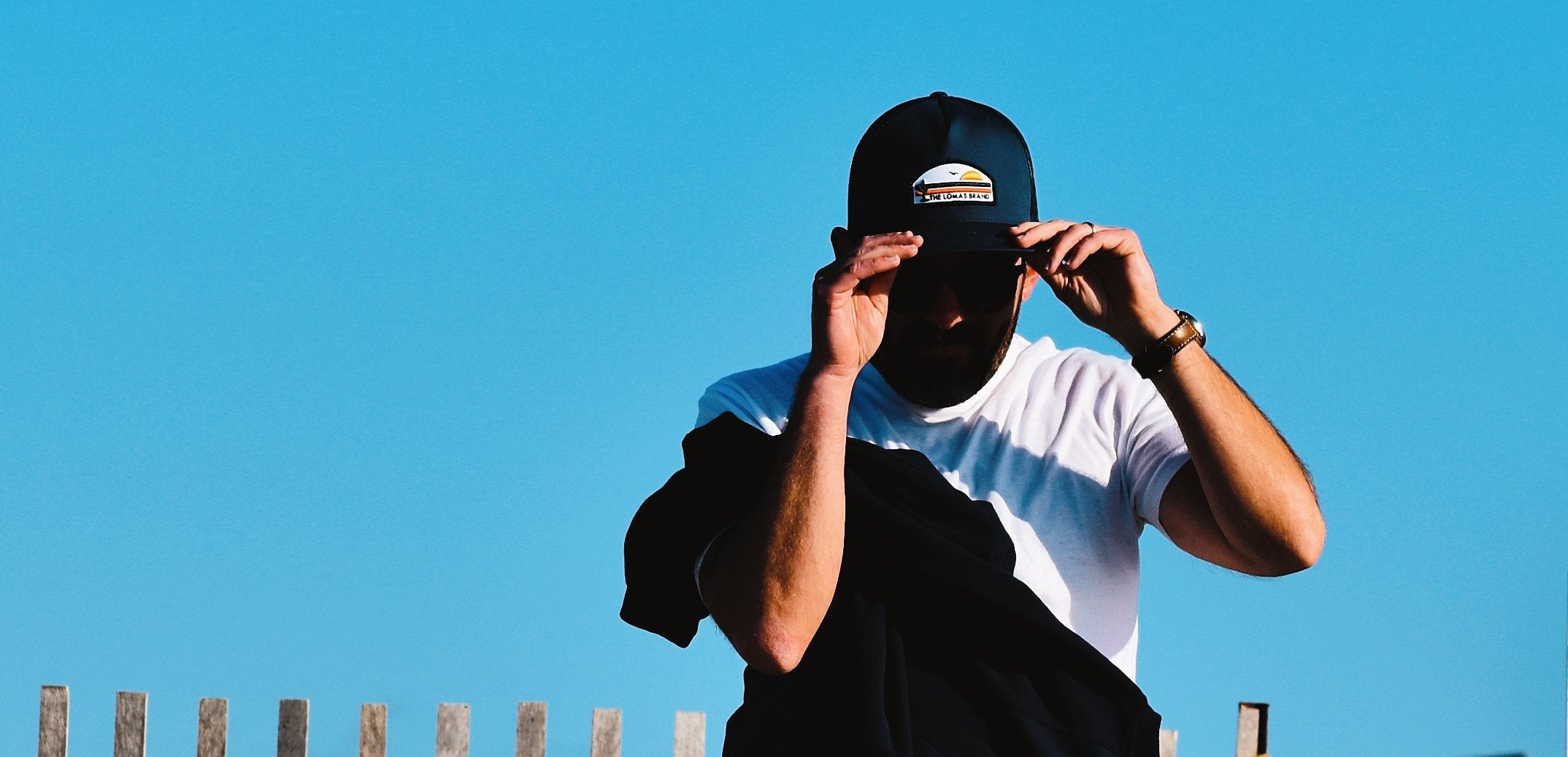 High contrast photo of a man fixing the brim of his navy blue hat with The Lomas Brand's colorful patch on the front. The man is wearing a white shirt and carrying a black jacket. The backdrop is the blue sky. 