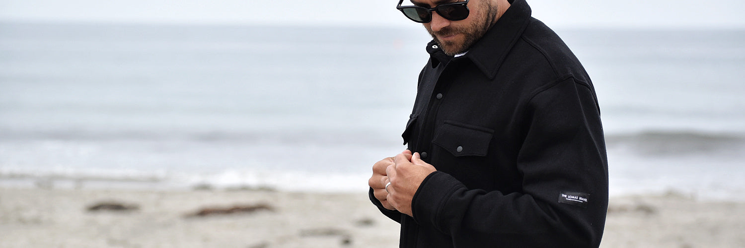 Mid body shot of a bearded man with arms crossed, smiling and looking at the surfboard next to him. The man is wearing a black t-shirt with The Lomas Brand design in white on the upper left chest. The vibe is happy. 