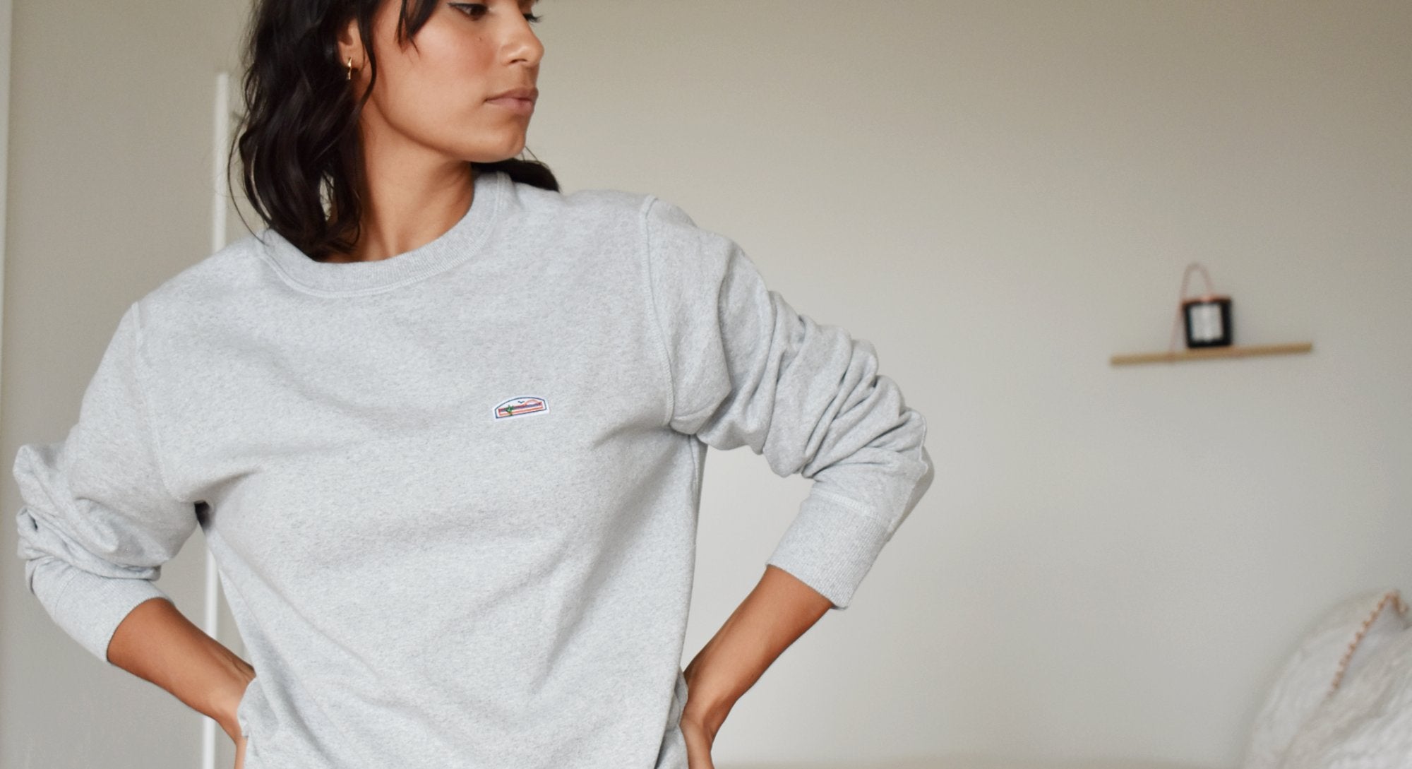 Woman with short dark hair wearing a grey crewneck sweatshirt with the sleeves pulled up. The sweatshirt features a small patch with The Lomas Brand logo on the upper left chest. The woman looks to the side and has her hands on her hips. 