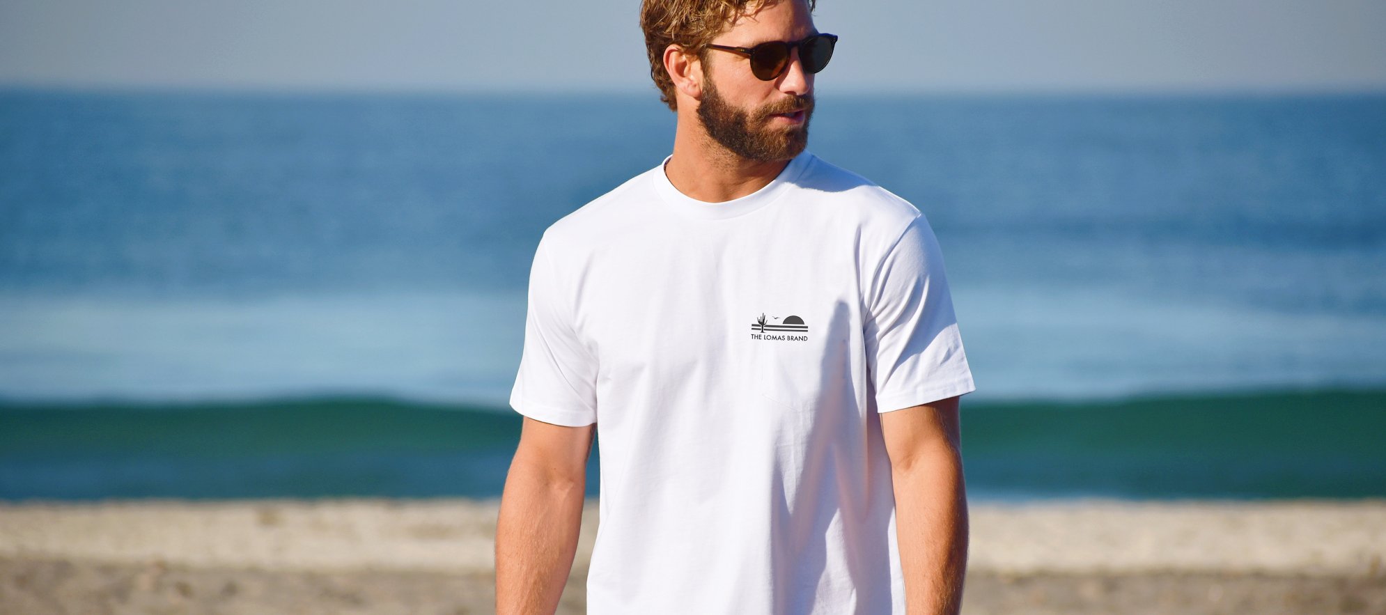 Man with sunglasses staring to the right of the camera wearing a white t-shirt with The Lomas Brand logo on the upper left chest, in black. The logo features a saguaro cactus, a rising sun, and a flying bird. The backdrop of the photo is the ocean.