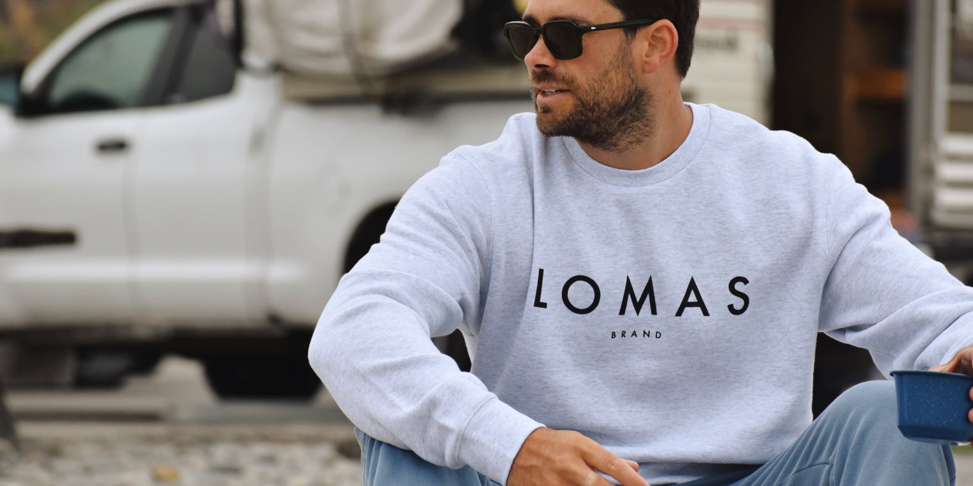 Man with sunglasses staring to the right of the camera wearing a white t-shirt with The Lomas Brand logo on the upper left chest, in black. The logo features a saguaro cactus, a rising sun, and a flying bird. The backdrop of the photo is the ocean.