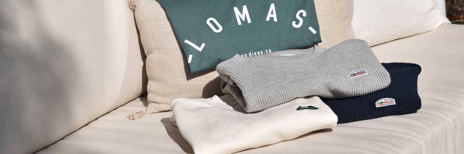 Smiling blonde woman with sunglasses faces at an angle to the camera. She is wearing a black bodysuit with a small white 'Lomas' written in a cursive script on the upper left chest.