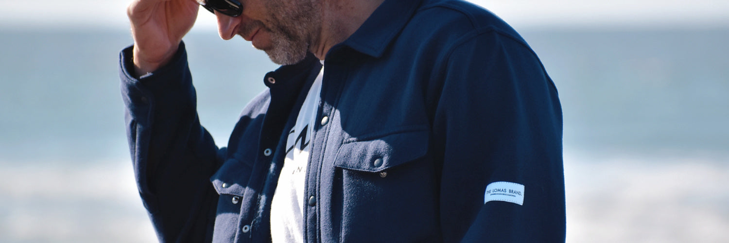 Photo of a man wearing sunglasses and a white shirt, putting on a dark crewneck sweatshirt. He stands near a parked car with the beach in the background. 
