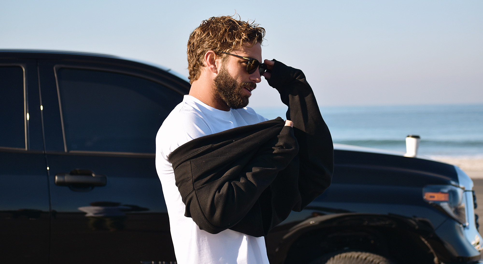 Photo of a man wearing sunglasses and a white shirt, putting on a dark crewneck sweatshirt. He stands near a parked car with the beach in the background. 