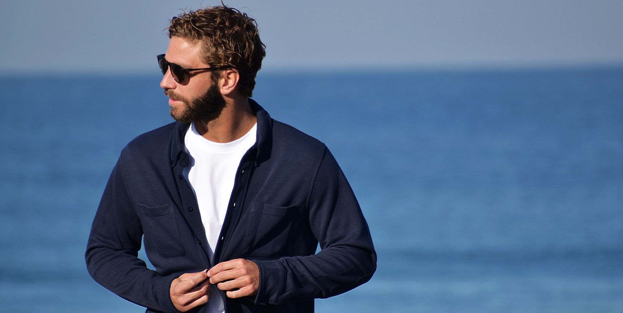 Man with beard and sunglasses at seaside, wearing navy button-up shirt over white tee. He's buttoning the shirt, looking to the side. Blue ocean and sky in background. Casual, stylish coastal look.