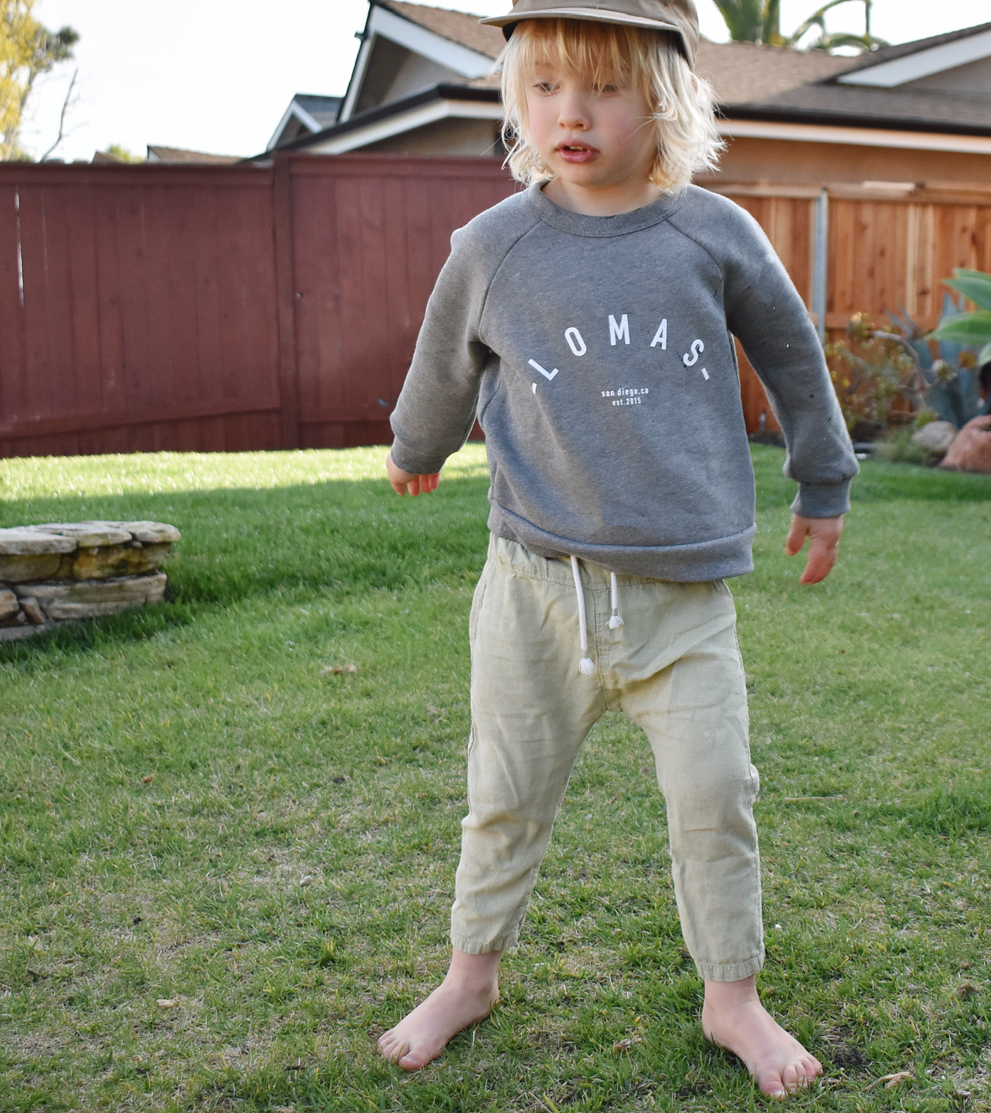 Toddler standing on some grass wearing a grey crew neck sweatshirt. The sweatshirt has a sweeping white arc that forms the text 'Lomas.' Situated beneath the arched logo are the phrases 'San Diego, CA' and 'est. 2015', denoting the location and establishment date.