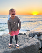 Laughing toddler on the rocks near the beach wearing a grey crew neck sweatshirt. The sweatshirt has a white arch shaped 'Lomas' written in all caps, 'San Diego, CA' 'est. 2015' under the arch.