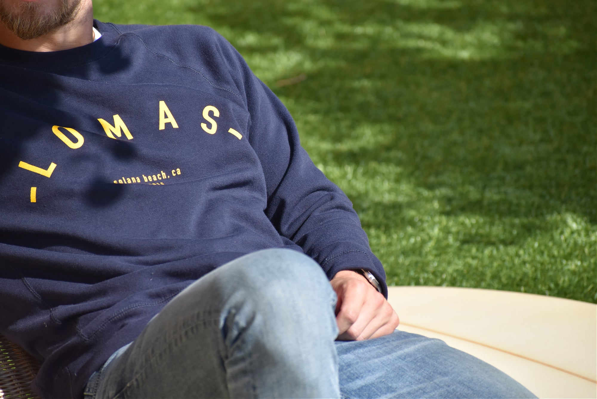 Close-up of someone wearing a navy blue sweatshirt with "LOMAS" printed in yellow letters across the chest. Below that, it says "solana beach, ca" in smaller yellow text. The person is sitting on what appears to be a wooden bench or surface, with green grass visible in the background.