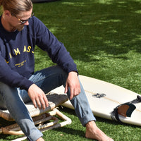 Man sits on chair in grass next to a surfboard wearing a navy crewneck sweatshirt with yellow/gold lettering. The Lomas Brand