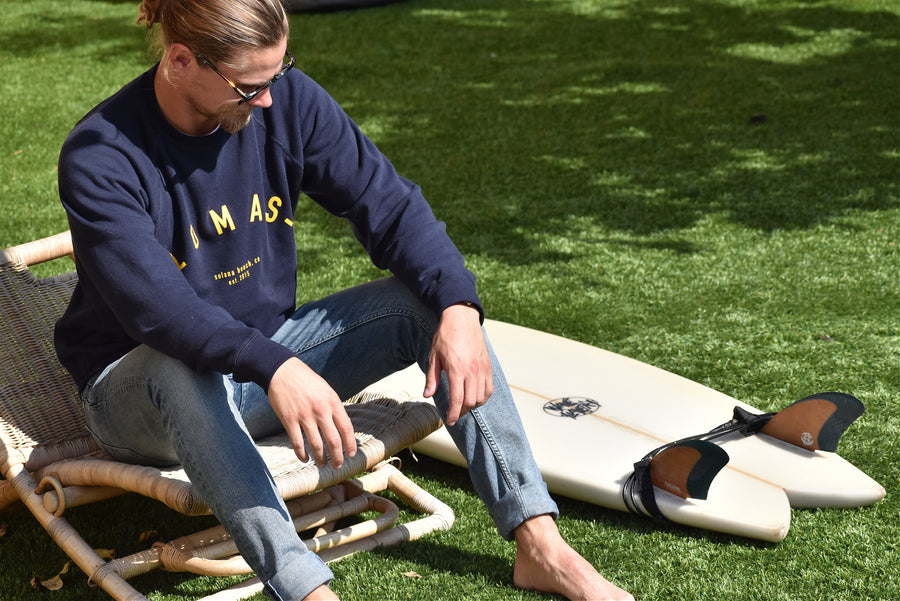 Man sits on chair in grass next to a surfboard wearing a navy crewneck sweatshirt with yellow/gold lettering. The Lomas Brand