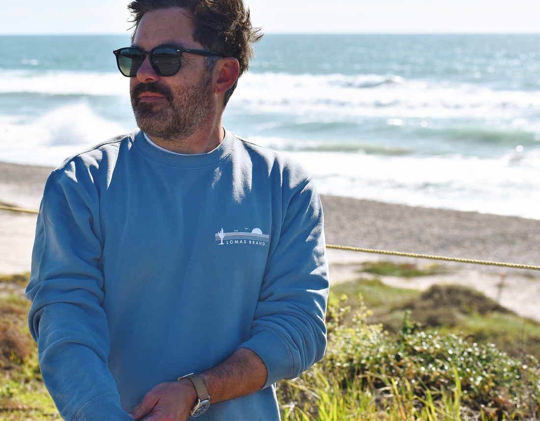 Upper body shot of a man with sunglasses standing in a tall grassy area with a beach in the background. The man is wearing a faded blue crewneck sweatshirt. The upper left of the sweatshirt features The Lomas Brand saguaro logo in white. The logo has a saguaro cactus, bird, and a rising sun in white. 'The' is printed above the logo and 'Lomas Brand' is printed below.
