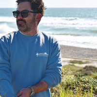 Upper body shot of a man with sunglasses standing in a tall grassy area with a beach in the background. The man is wearing a faded blue crewneck sweatshirt. The upper left of the sweatshirt features The Lomas Brand saguaro logo in white. The logo has a saguaro cactus, bird, and a rising sun in white. 'The' is printed above the logo and 'Lomas Brand' is printed below.