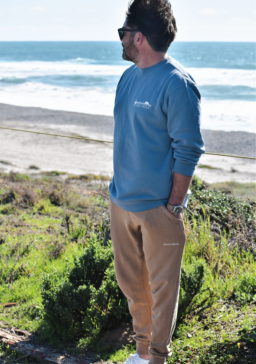 A full-length photo capturing a man wearing sunglasses with his gaze averted from the camera. He is standing in a tall grassy area with a beach visible in the background. The man's outfit consists of a faded blue crewneck sweatshirt and camel brown sweatpants. On the upper left of the sweatshirt, an emblematic logo in white depicts a stylized saguaro cactus with outstretched limbs, a soaring a bird, and an ascending sun. Above the emblem is the word 'The,' with 'Lomas Brand' below the graphic elements.