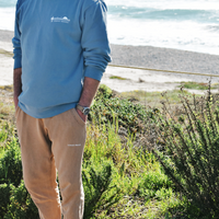 Mid-body photo of a man standing in a tall grassy area with a beach in the background. The man is wearing a faded blue crewneck sweatshirt and camel brown sweatpants. The upper left of the sweatshirt features The Lomas Brand saguaro logo in white. The logo has a saguaro cactus, bird, and a rising sun in white. 'The' is printed above the logo and 'Lomas Brand' is printed below.