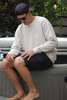 Man sitting on the edge of a hot tub or small pool. He's wearing a beige crewneck sweatshirt with a small chest logo, black shorts, sunglasses, and a backwards black cap. The setting looks like an outdoor patio area with plants and a hanging light bulb visible in the background.