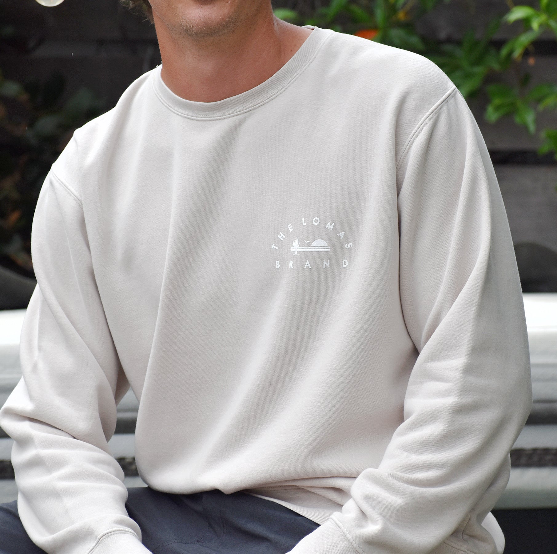 Person wearing a beige crewneck sweatshirt. The sweatshirt has a small logo on the chest that reads "THE LOMAS BRAND" with a small icon. The icon shows a saguaro cactus, a bird, and the rising sun. The person is shown from the neck down, seated outdoors with some greenery visible in the background.