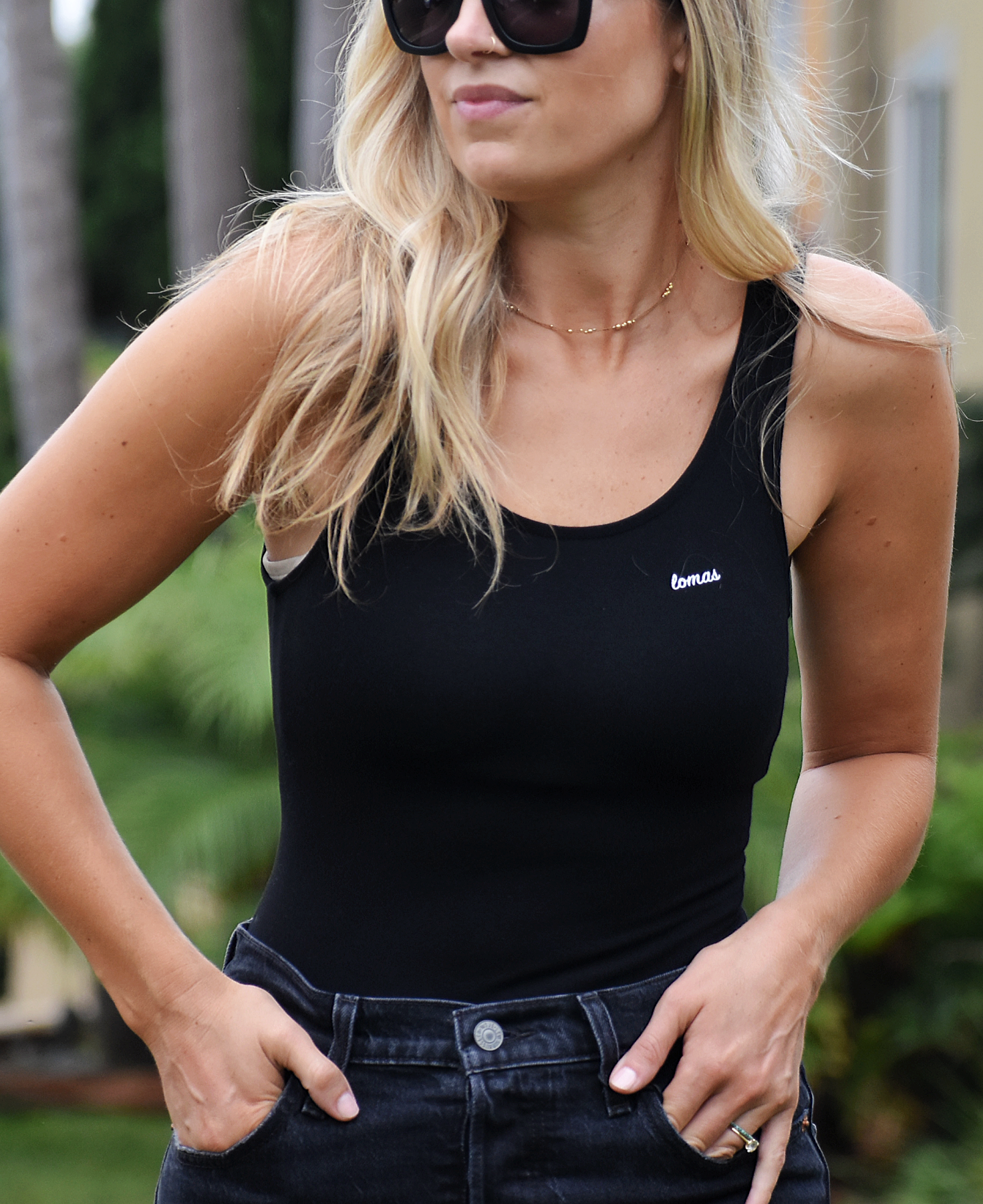 A blonde woman wearing a black tank top bodysuit with a small logo on the chest, paired with high-waisted black jeans. She is adjusting her jeans while standing outdoors.