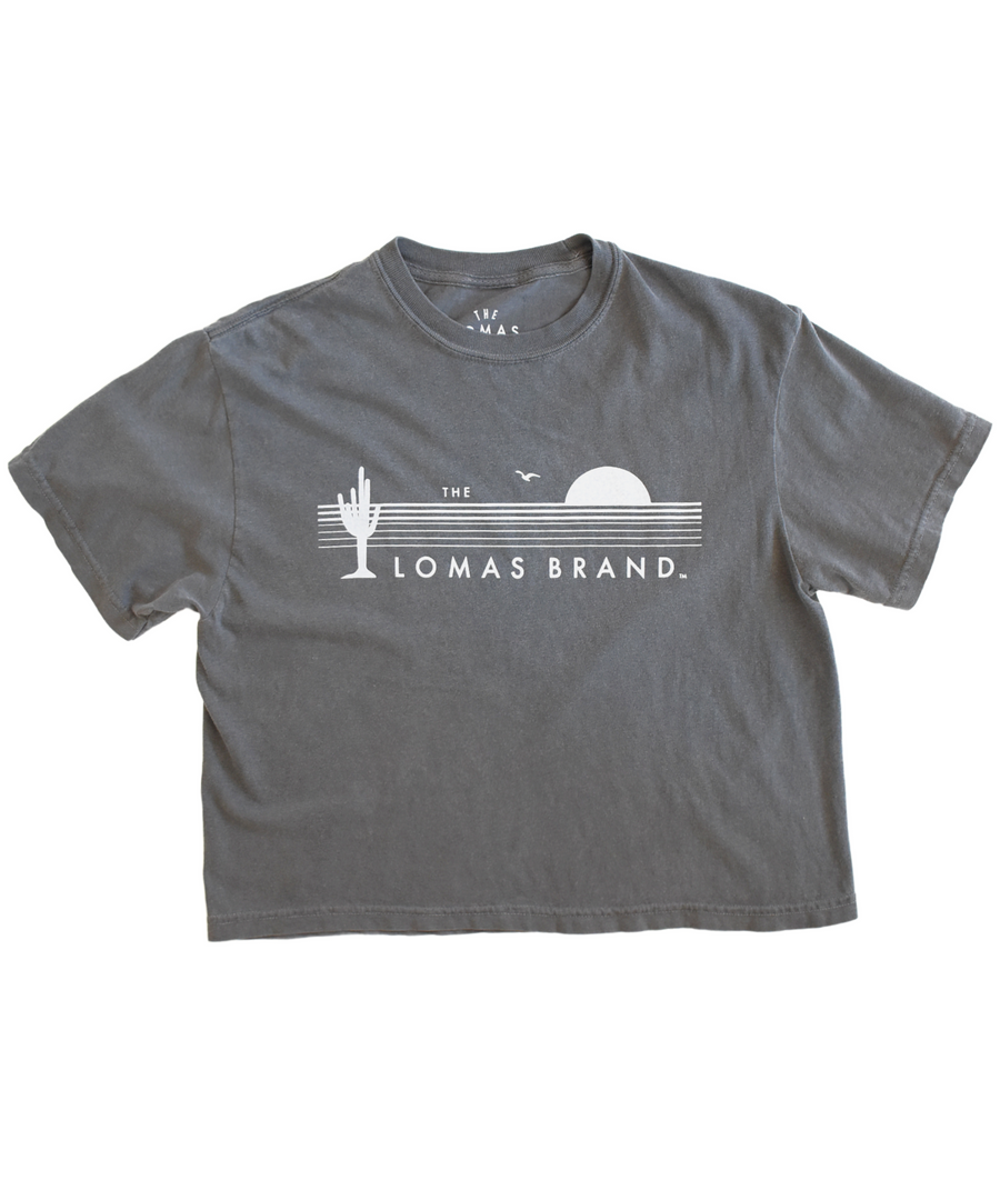 Premium heavy cotton grey t-shirt with a shorter fit just above the waist. The shirt bears an emblem rendering a saguaro cactus with raised stems, a soaring bird, and a dawning sun in a rich white hue. The word 'The' falls just above the graphic. Below the logo, the words 'Lomas Brand' is proudly shown.