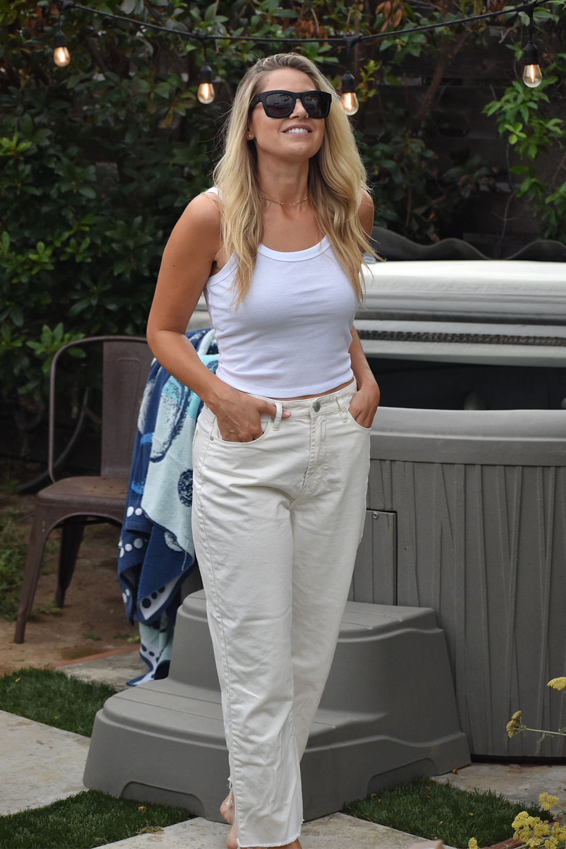 Smiling blonde woman with sunglasses stands near a hot tub wearing a white cami crop top and white jeans. Her hands are in her pockets.