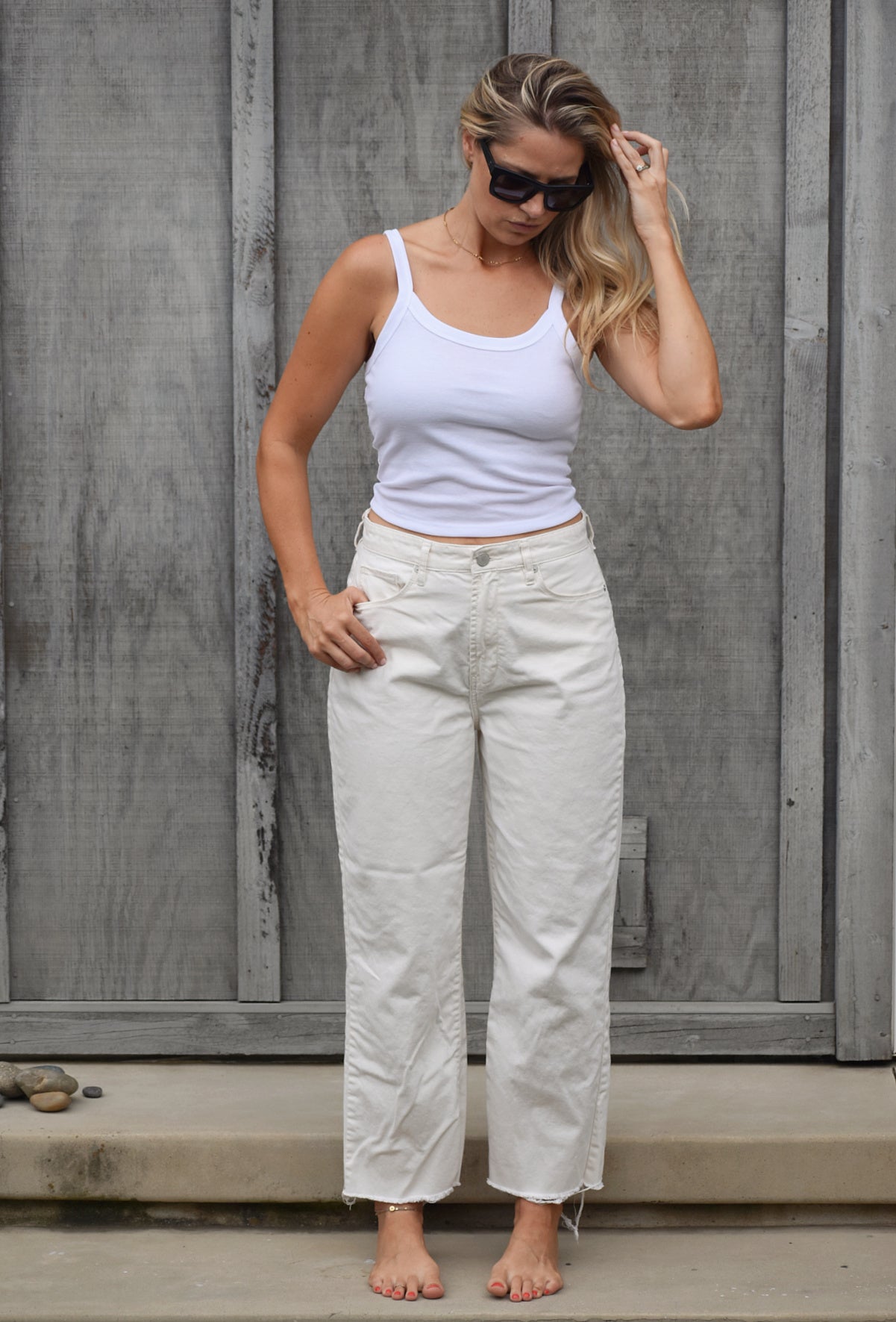 Blonde woman runs her hand through her hair while looking down. She is wearing a white cami crop top tank and white jeans. She stands barefoot on steps with a wood backdrop.