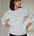 Woman wearing a grey crewneck with her hands on her hips looking off camera. The sleeves are rolled up and the sweatshirt looks cozy. There is a small patch on the upper left chest with a saguaro cactus, a bird, and a rising sun. 