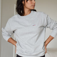 Woman wearing a grey crewneck with her hands on her hips looking off camera. The sleeves are rolled up and the sweatshirt looks cozy. There is a small patch on the upper left chest with a saguaro cactus, a bird, and a rising sun. 