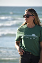 A photo of a woman wearing sunglasses and standing on a beach, staring off to the left side of the frame. She is dressed in a green t-shirt and black jeans. The t-shirt features a graphic depicting a saguaro cactus with outstretched arms, a soaring bird, and a radiant sun below, all rendered in white. The word 'The' appears above the emblem, while 'Lomas Brand' is inscribed underneath the graphic elements. A beach setting provides the backdrop for the image.