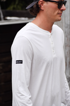 Close-up side view of a man wearing a white henley shirt. It shows the details of the shirt, with a small brand tag on the sleeve that reads "The Lomas Brand."