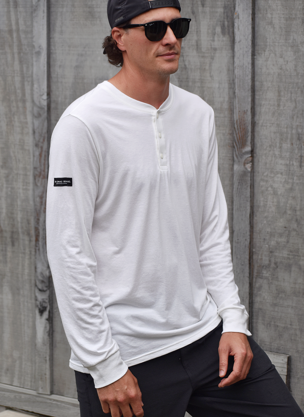 Man facing towards the camera. He's wearing a white long-sleeved henley shirt, sunglasses, and backwards cap. The background shows wooden planks, suggesting an outdoor setting.