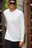 Man wearing a white long-sleeved henley shirt, dark sunglasses, and a backwards baseball cap. He is standing in what looks like a doorway or entrance, with wooden structures visible in the background. The man has short, curly hair visible under his cap.