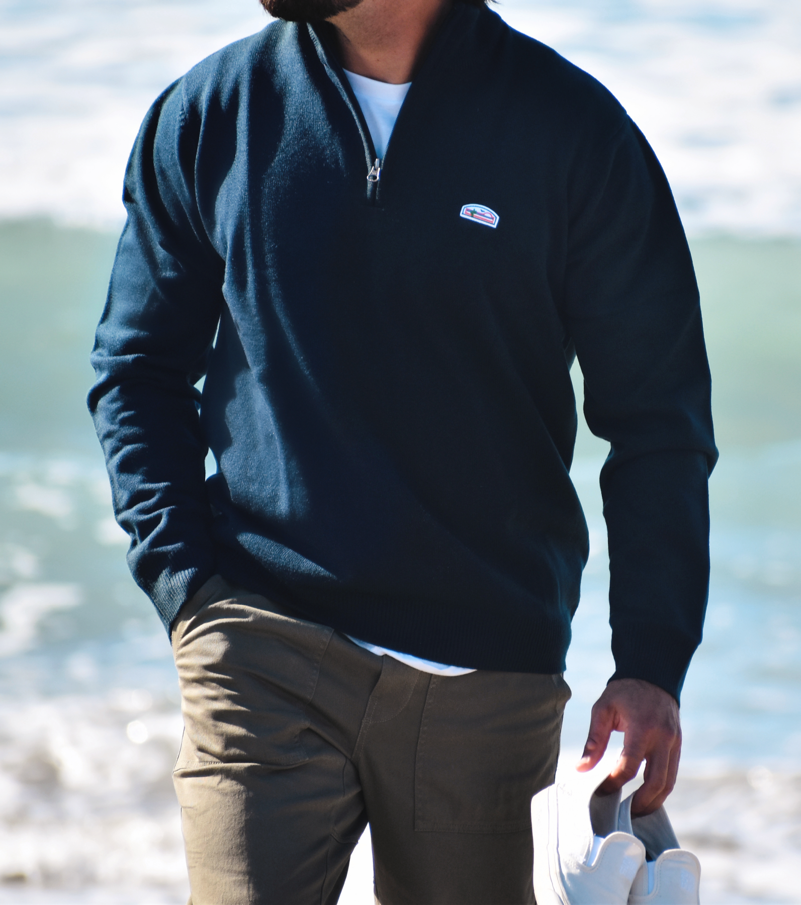 A mid-body photo of a man wearing a comfortable and warm navy quarter zip with a white t-shirt underneath and khaki pants. He is carrying white slip on shoes with one hand and has his other hand in his pocket. Gracing the upper left side of the quarter zip is The Lomas Brand's emblem in patch form, showcasing a saguaro cactus, a bird taking flight, and an ascending sun – all brought to life in vibrant white, orange, and green hues.