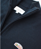 Close-up of a navy blue sweater or pullover, focusing on the embroidered logo patch. The patch shows a stylized sun, mountains, and cactus design with "THE LOMAS BRAND" text underneath.