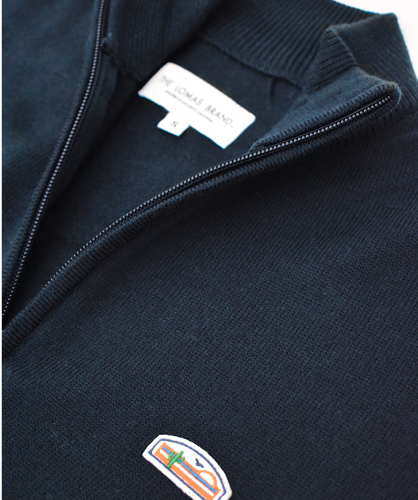 Close-up of a navy blue sweater or pullover, focusing on the embroidered logo patch. The patch shows a stylized sun, mountains, and cactus design with "THE LOMAS BRAND" text underneath.