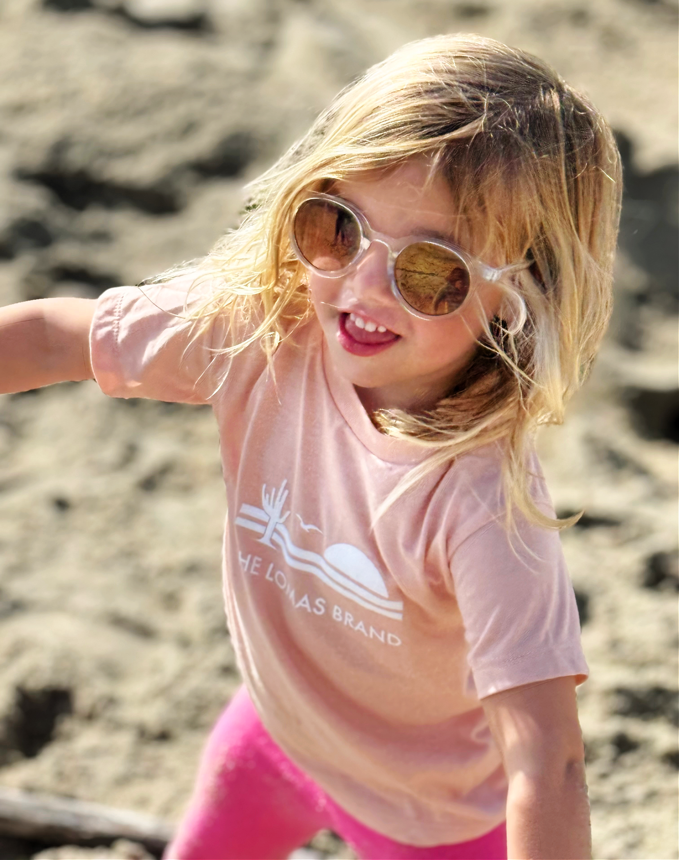 Smiling toddler on the beach with sunglasses wearing a soft pink t-shirt. The shirt has The Lomas Brand logo with a saguaro cactus, bird, and a rising sun in white. 'The Lomas Brand' is printed below.