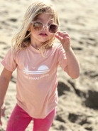 Toddler on the beach with sunglasses wearing a soft pink t-shirt. The shirt has The Lomas Brand logo with a saguaro cactus, bird, and a rising sun in white. 'The Lomas Brand' is printed below.