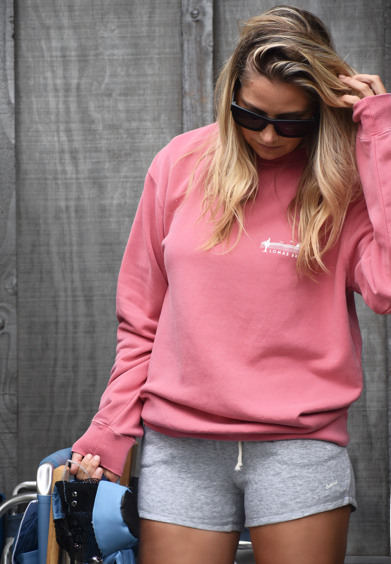 A woman with long blonde hair wearing sunglasses and a salmon colored sweatshirt. She is running her hand through her hair and wearing grey shorts. A wooden fence is visible in the background.