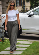 A woman walking on a paved path with grass and plants visible around her. A white car can be seen in the background. She's wearing a white t-shirt, olive green pants, and sunglasses. Her blonde hair is flowing in the wind.