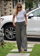 A woman walking on a path next to a grassy area. A white car and some garden equipment are visible. She's wearing a white t-shirt, olive pants, and sunglasses.